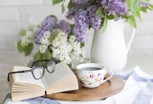 Coffee, a book and reading glasses, and a white vase with purple and white lilacs on a table top.