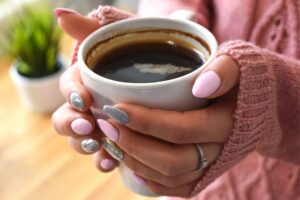 a woman's hands - with pink and gray fingernail polish - partly covered by pink sweater sleeves - holds a cup of coffee that has left a ring in the coffee cup from being out too long before she could drink.
