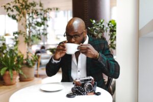 A man drinking coffee at a round table outdoors. His camera is on the tablecloth. 